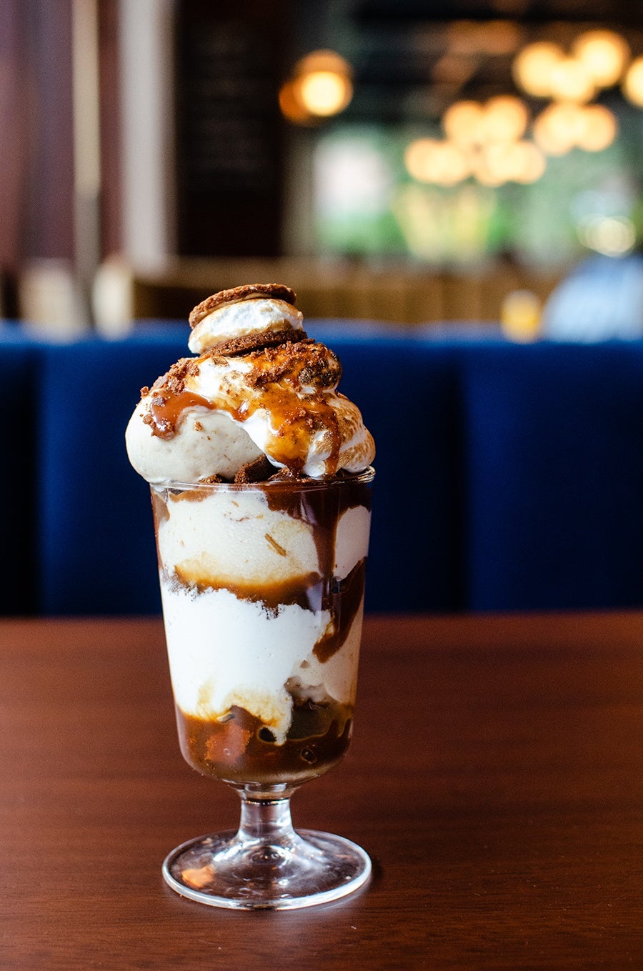 A tall glass filled with a layered dessert featuring vanilla ice cream, caramel sauce, and chocolate syrup, topped with whipped cream and a small cookie. The dessert is placed on a wooden table with a blurred background of a blue cushioned bench and warm ambient lights.