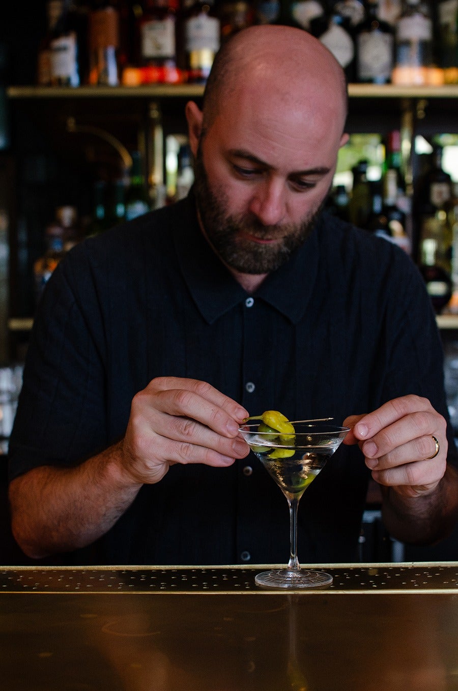 A bald man with a beard, wearing a dark button-up shirt, is carefully placing a garnish of green peppers on a martini glass filled with a clear liquid, standing behind a bar counter with bottles blurred in the background.