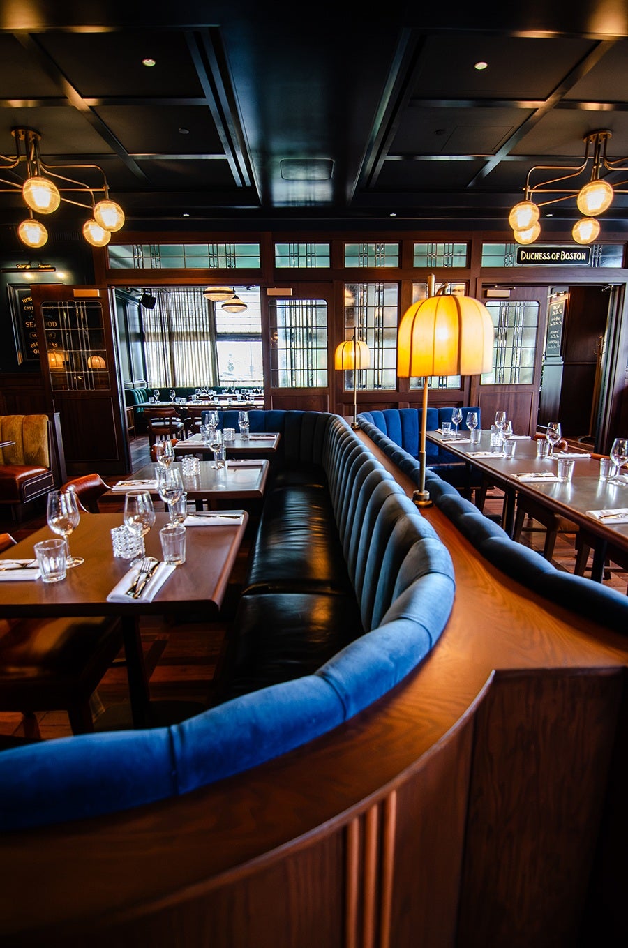A stylish restaurant interior featuring a curved booth with blue velvet upholstery and black leather seating. The booth is accompanied by wooden tables set with glassware, napkins, and cutlery. Warm yellow lamps with dome-shaped shades are mounted on the booth's wooden divider. The background shows wooden paneling, glass windows with geometric patterns, and additional dining tables and chairs. The ceiling is dark with recessed lighting and modern chandeliers with round bulbs. A sign reading "Duchess of Boston" is visible above a doorway.