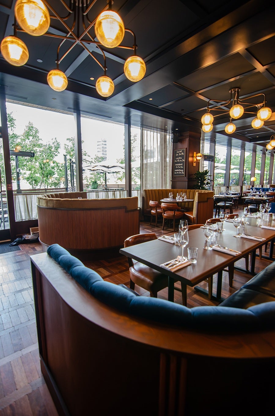 Modern restaurant interior with curved wooden booths upholstered in blue and tan fabric, dark wood tables set with glassware and napkins, large floor-to-ceiling windows allowing natural light, and warm pendant lighting fixtures hanging from a dark ceiling.