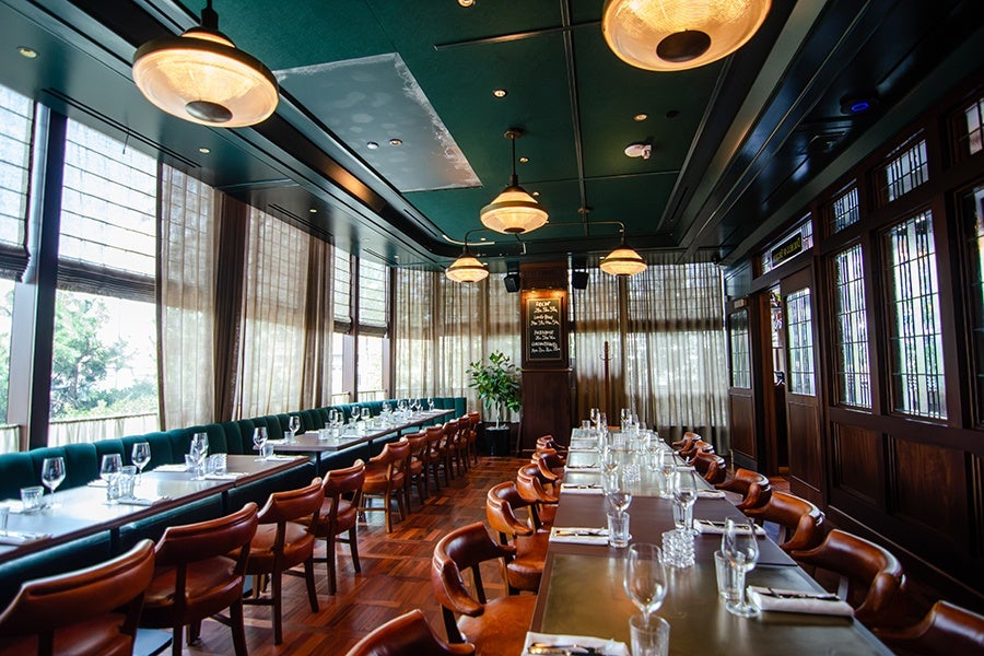 A spacious, elegant dining room with long tables set for a meal. The tables have white napkins, glassware, and cutlery neatly arranged. The room features dark wood paneling, large windows with sheer curtains, green upholstered bench seating along one side, and brown leather chairs. Warm pendant lights hang from a dark green ceiling, and a chalkboard menu is visible at the far end near a potted plant. The floor is polished wood parquet.
