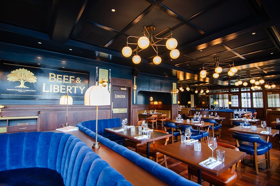 Upscale restaurant interior with dark wood paneling, blue velvet curved banquette seating, wooden tables set with glassware and napkins, and modern globe pendant lighting fixtures hanging from a dark coffered ceiling. A large sign on the wall reads "Beef & Liberty.