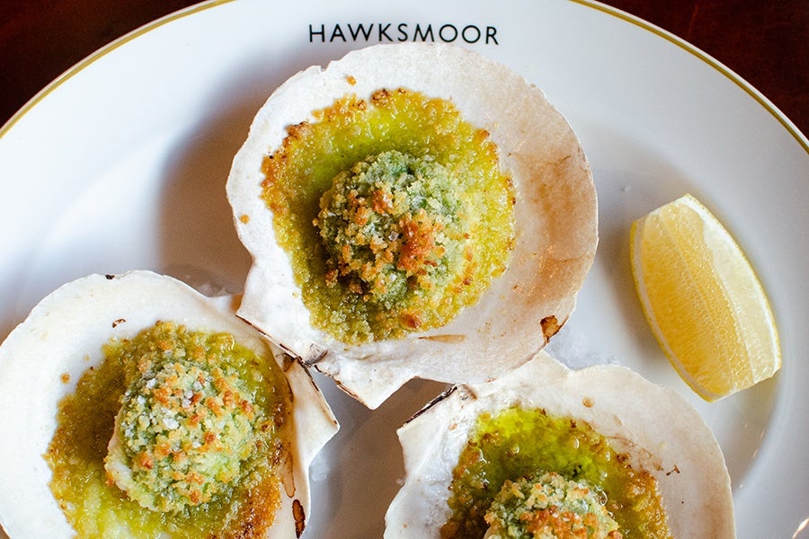 Three baked scallops in shells topped with a green herb and breadcrumb crust, served on a white plate with a lemon wedge. The plate has the word "HAWKSMOOR" printed at the top.