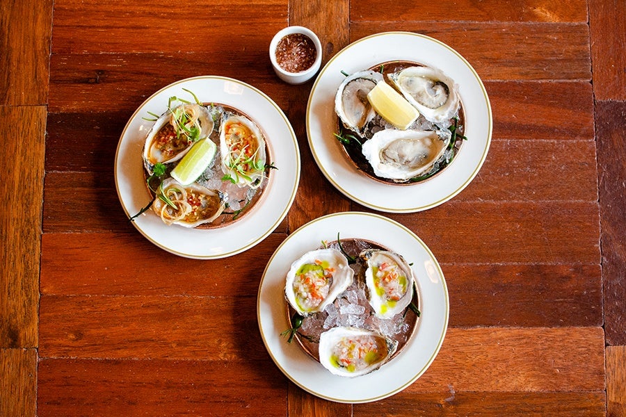 Three plates of oysters on the half shell, each served on a bed of ice with garnishes. One plate has oysters topped with a red sauce and a lime wedge, another has plain oysters with a lemon wedge, and the third has oysters with a diced vegetable topping and a drizzle of green sauce. A small cup of coarse salt is placed above the plates on a wooden table.