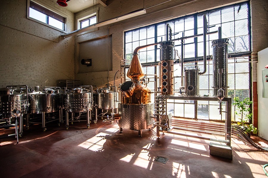 A distillery setup with a large copper and stainless steel still in the center, surrounded by multiple stainless steel fermentation tanks on the left. The room has high ceilings, large industrial windows letting in natural light, and a red concrete floor. Pipes and metal structures connect the equipment.