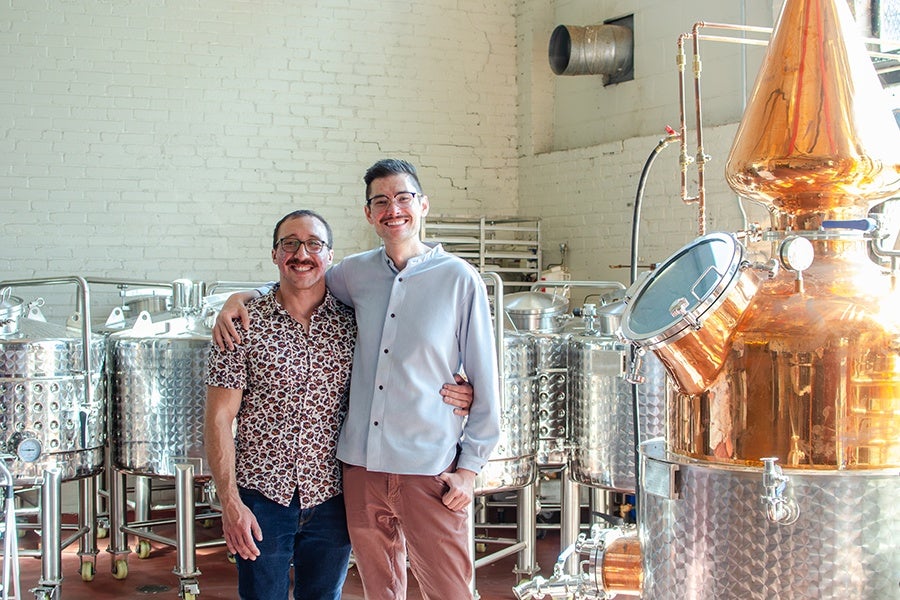 Two men standing side by side with their arms around each other in a distillery. They are smiling and wearing glasses; one is in a patterned short-sleeve shirt and jeans, the other in a light-colored button-up shirt and brown pants. Behind them are several stainless steel fermentation tanks and a large copper distillation apparatus. The setting is an industrial space with white brick walls.