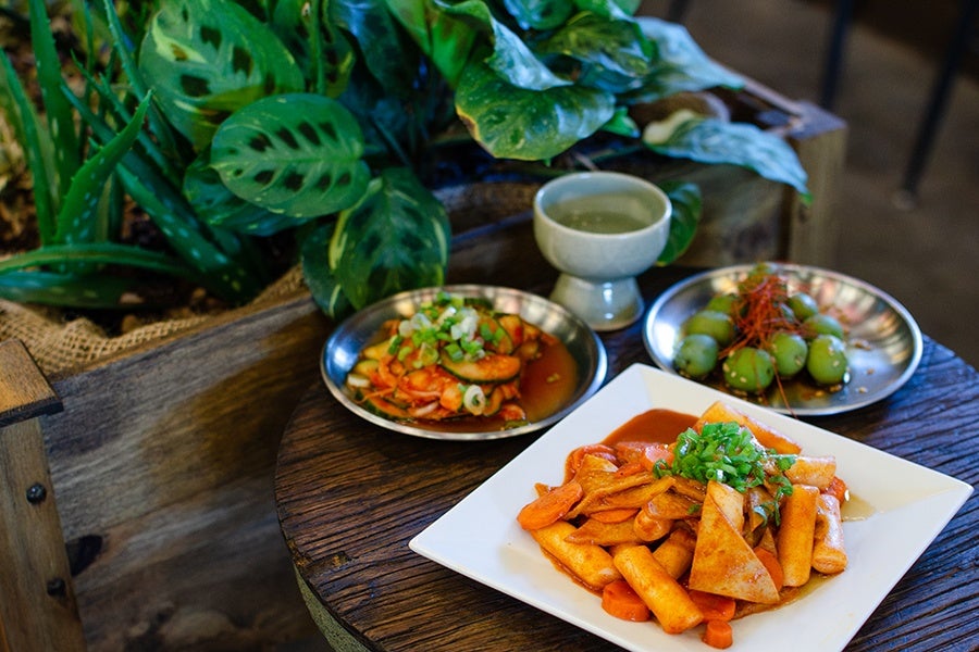 A wooden table holds three dishes and a cup. The main dish on a white square plate contains stir-fried rice cakes with sliced carrots and green onions in a reddish sauce. Behind it, a metal plate has green olives topped with thin red strands, and another metal plate holds a shrimp dish garnished with chopped green onions. A small ceramic cup filled with a light-colored liquid is also on the table. The background features green leafy plants and wooden surfaces.