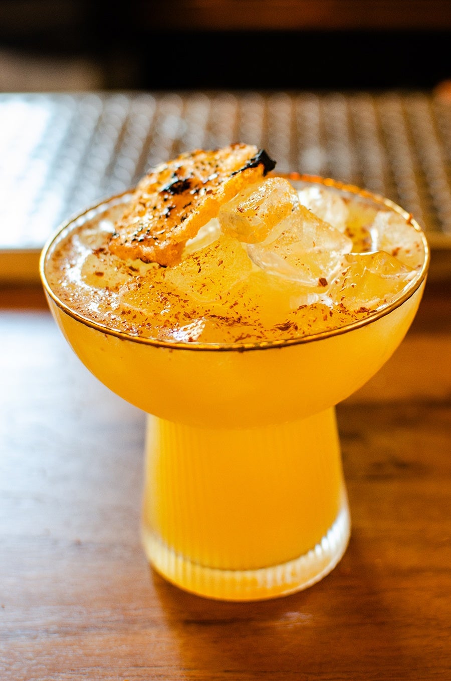 Yellow-orange cocktail served in a wide, shallow glass with a gold rim, filled with ice cubes and garnished with a charred citrus slice and a sprinkle of spices. The glass has a textured, ribbed base and is placed on a wooden surface.
