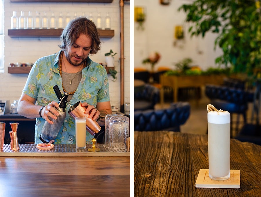 A man with shoulder-length hair and a beard, wearing a light blue floral shirt and a beaded necklace, is pouring a drink from two metal shakers into a tall glass on a bar counter. The bar has copper measuring tools and a sliced grapefruit. On the right, a tall frothy cocktail garnished with a dried citrus slice sits on a wooden coaster on a rustic wooden table, with a blurred background of a cozy seating area featuring dark blue tufted chairs and green plants.