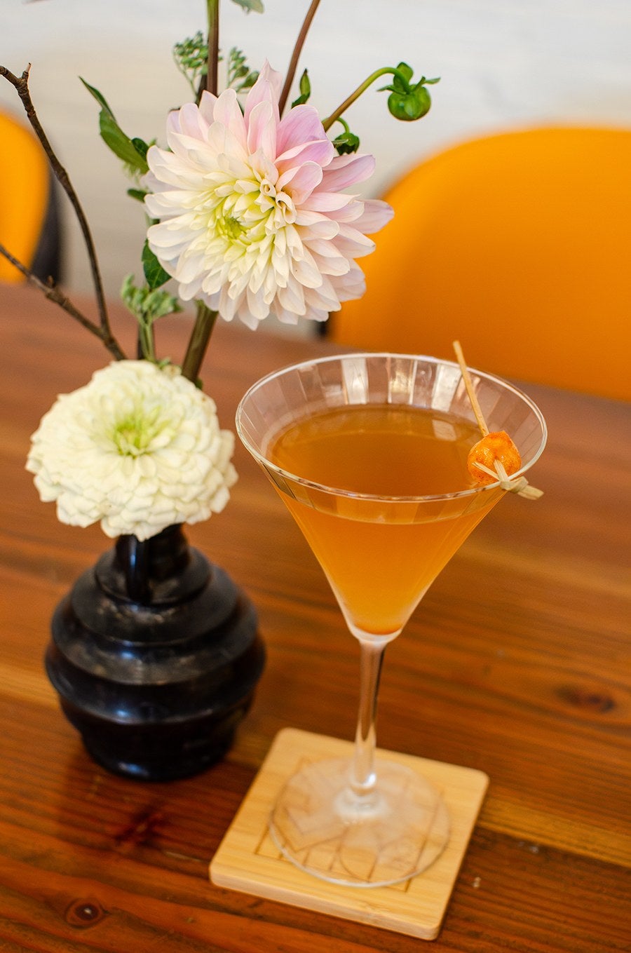 A clear martini glass filled with an amber-colored cocktail, garnished with a small orange peel twist on a wooden skewer, placed on a square wooden coaster. Next to the glass is a black vase holding two large flowers, one white and one pale pink with green centers, along with some green foliage. The setting is on a wooden table with an orange chair in the background.