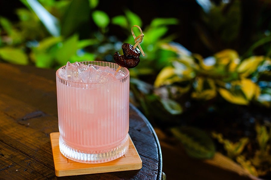 A pink cocktail served in a clear, ribbed glass filled with crushed ice, garnished with two dark cherries on a wooden pick, placed on a square wooden coaster on a dark wooden table with green foliage in the background.