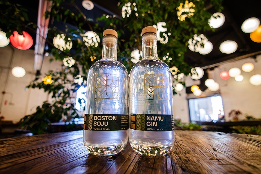 Two clear glass bottles with wooden caps are placed on a wooden surface. The bottle on the left is labeled "Boston Soju" and the one on the right is labeled "Namu Gin," both with black and gold labels indicating 40% alcohol by volume. The background features blurred hanging lights and greenery, creating a warm, ambient setting.