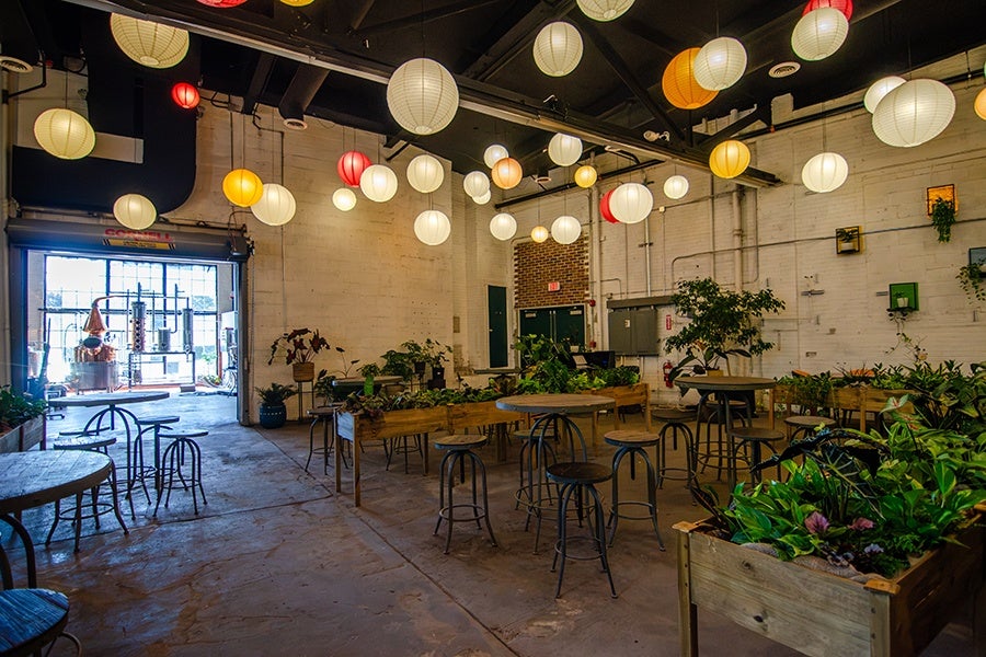 A spacious indoor area with concrete floors and white brick walls, decorated with numerous hanging paper lanterns in white, yellow, and red. The room features several round tables with metal stools and raised wooden planters filled with green plants. Large windows and an open garage-style door allow natural light to enter. The ceiling is black with exposed beams and pipes.