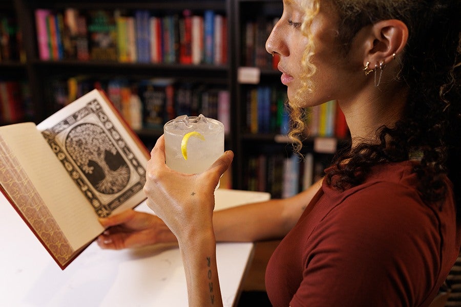 A person with curly hair and multiple earrings is holding a glass of iced lemon drink in one hand and an open book with an intricate black and white illustration of a tree in the other hand. They are seated at a white table with a bookshelf filled with colorful books in the background.