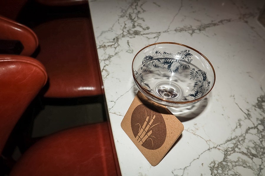 A clear glass bowl with black leaf patterns and a gold rim is placed on a cork coaster featuring a bamboo design. The bowl and coaster are on a white marble surface with gray veining. Red leather chairs are partially visible in the background.