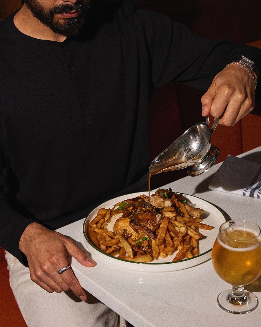A person wearing a black long-sleeve shirt is pouring gravy from a silver gravy boat onto a plate of roasted chicken and French fries garnished with herbs. A glass of beer is placed on the table next to the plate.