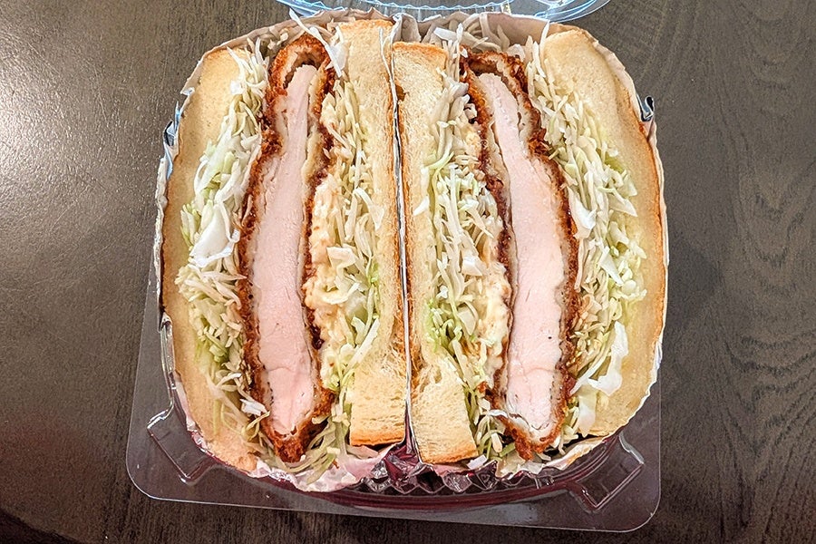 Two sandwich halves with breaded chicken cutlets, shredded cabbage, and a creamy sauce, placed in a clear plastic container on a wooden surface.